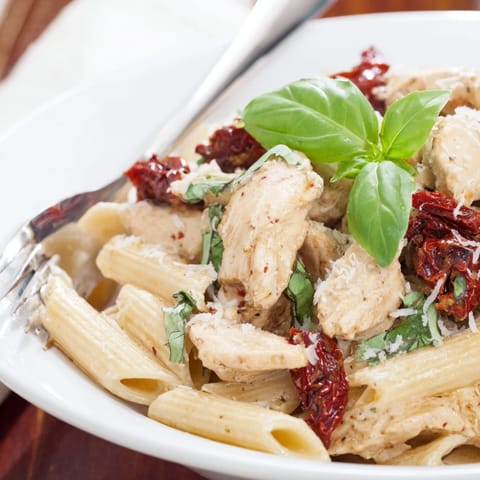 A close-up of Creamy Sun-Dried Tomato Chicken Pasta, highlighting glossy sauce clinging to penne with chicken and sun-dried tomatoes, garnished with fresh basil.  